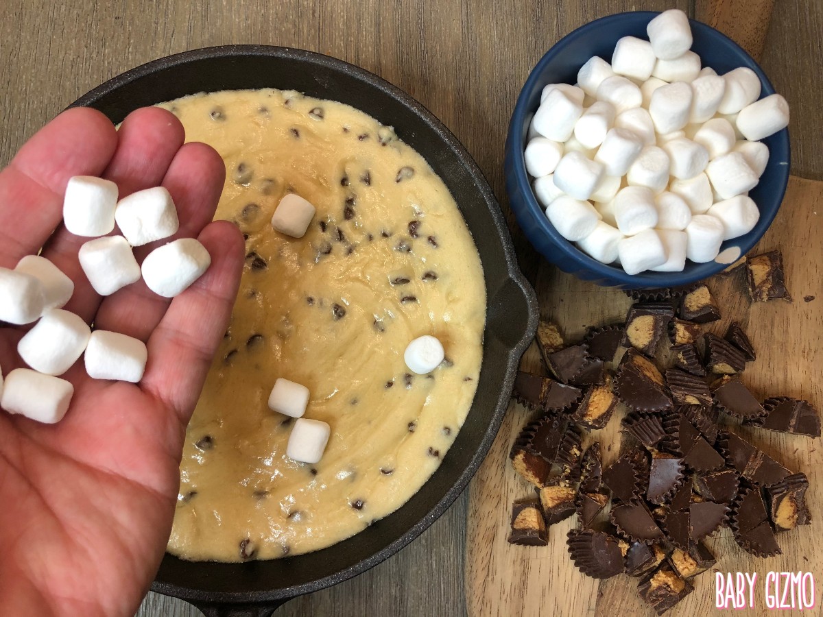 cookie dough in skillet with handful of mini marshmallows and chopped peanut butter chocolate candies