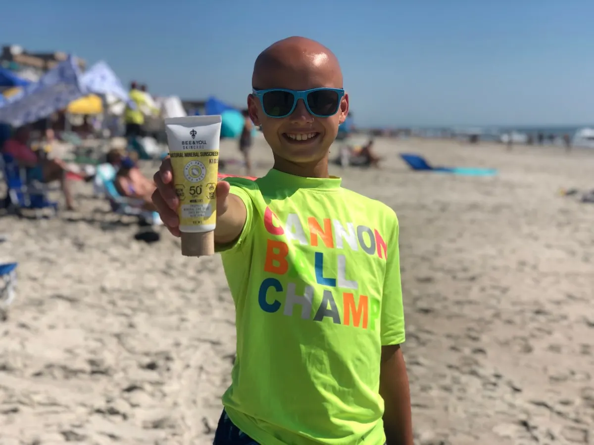 bald boy holding out sunscreen in hand on the beach