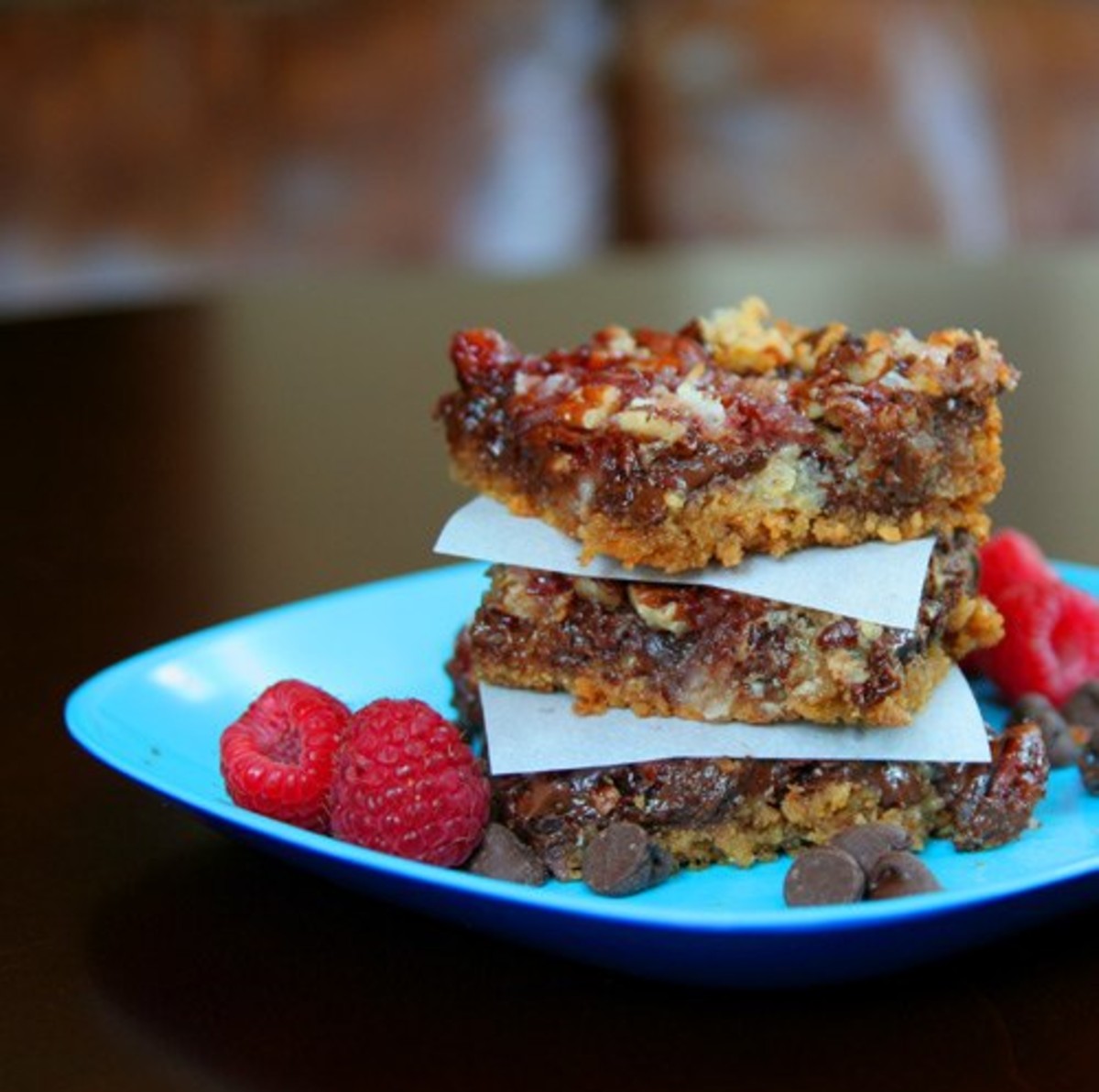 3 raspberry magic bars stacked on top of each other on a blue plate with raspberries and chocolate chips on the side  